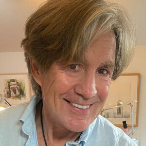 A smiling middle-aged man with light brown hair and a blue shirt stands indoors, framed artwork and soft lighting behind him. He is the PleinAir Salon Art Competition Judge for March 2025. 15th Annual PleinAir Salon Art Competition