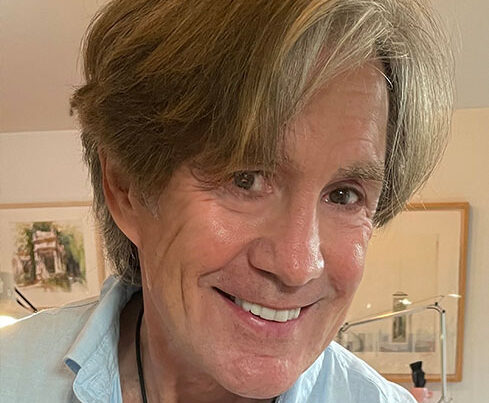 A smiling middle-aged man with light brown hair and a blue shirt stands indoors, framed artwork and soft lighting behind him. He is the PleinAir Salon Art Competition Judge for March 2025. 15th Annual PleinAir Salon Art Competition