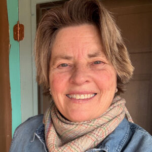 A smiling older woman with short, light brown hair wears a plaid scarf and denim jacket, standing indoors in front of a brown door and teal wall. She is the PleinAir Salon Art Competition Judge for March 2025. 15th Annual PleinAir Salon Art Competition