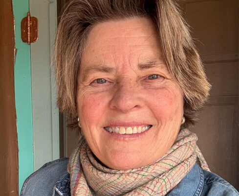 A smiling older woman with short, light brown hair wears a plaid scarf and denim jacket, standing indoors in front of a brown door and teal wall. She is the PleinAir Salon Art Competition Judge for March 2025. 15th Annual PleinAir Salon Art Competition